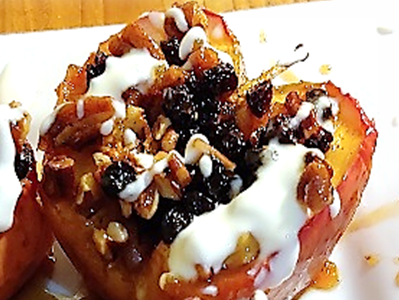 Baked apple covered in raisins, syrup, and creme fraiche, and pecans. The apple sits on a white plate atop a wooden surface.