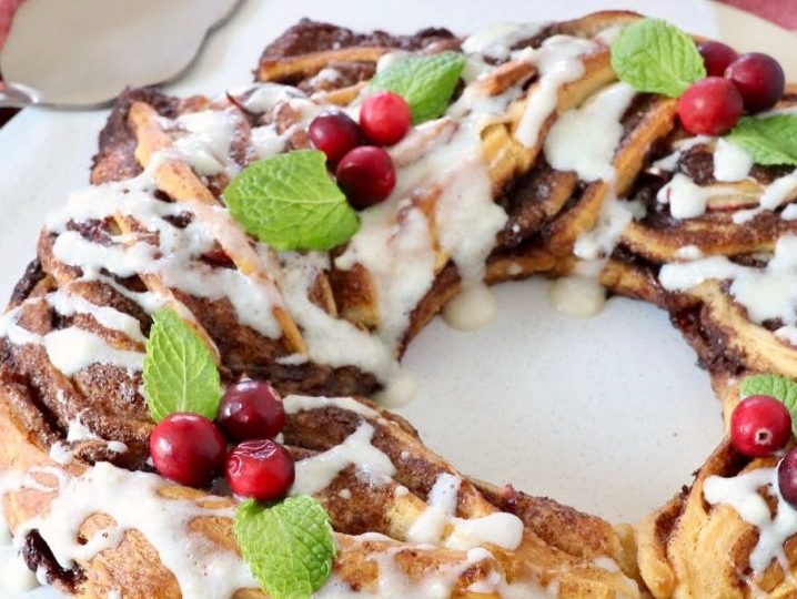 apple cinnamon pastry wreath that is adorned with cranberries and mint. Icing drips down the sides. This all sits on a white surface.