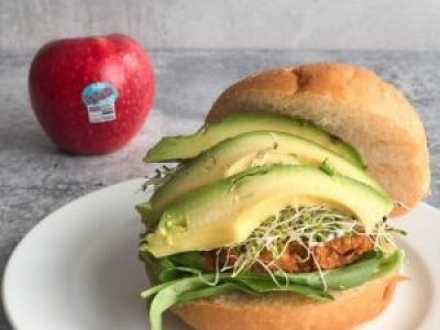 pazazz apple sitting behind a chickpea apple burger with avocado and sprouts spilling out from under the bun.