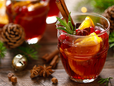 glass of red tinted Pazazz apple sangria with a stick of cinnamon and orange rind in the glass. Spices, greenery, bells, and pinecones adorn the table around the glass.